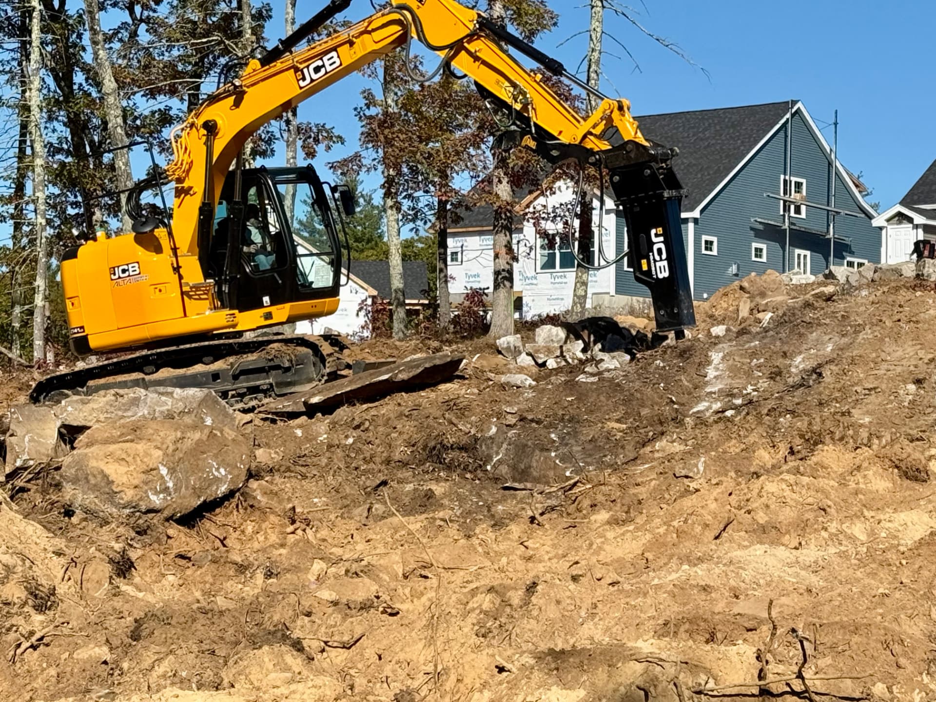 Yellow JCB excavator with a hydraulic breaker working a rocky New Hampshire job site